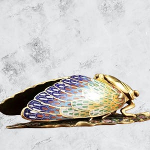 Op de afbeelding: Een decoratief cicadebeeldje van goudkleurig metaal met kleurrijke emaille vleugels. De vleugels hebben blauwe, groene en gele patronen. Het beeldje staat op een blad-vormige basis, met ingewikkelde details.