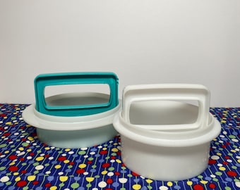 May include: Two white plastic containers with teal and white handles. The containers are round with a handle on top. The containers are on a blue patterned surface with red, yellow, and green dots.