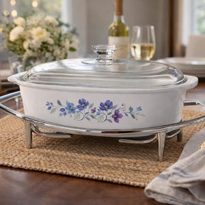 May include: White oval casserole dish with a glass lid, featuring a floral design in blue and purple. The dish sits on a silver-toned metal warming stand, perfect for serving. A candle, flowers, and wine glasses are in the background.