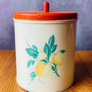 May include: Vintage cream-colored metal canister with a bright orange lid and knob. The canister features a floral design with yellow lemons, teal leaves, and white flowers. A charming kitchen storage container.
