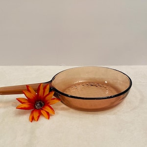 May include: A vintage amber-colored glass skillet with a long handle and a textured interior. The skillet has a black rim and is accompanied by a vibrant orange and yellow flower. A classic kitchenware item.