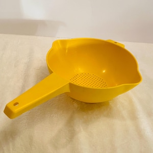 May include: A bright yellow plastic colander with a long handle and a perforated straining surface. The colander has a rounded bowl shape with a pouring spout and a hole at the end of the handle.
