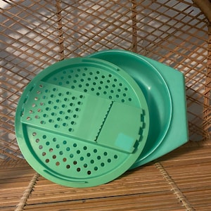 May include: A green plastic colander with a built-in grater. The colander has a round shape and a handle on the side. The grater is located on the top of the colander and has a rectangular shape.