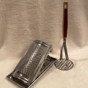 May include: A stainless steel grater and a potato masher with a wooden handle. The grater is rectangular with a tray, and the masher has a circular head. Both are kitchen tools.