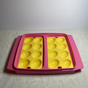 May include: A pink and yellow plastic deviled egg tray. The tray has a rectangular shape with a raised edge and two sections of yellow egg-shaped indentations. The tray is designed to hold a dozen deviled eggs.