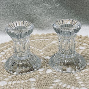 May include: Two clear glass candle holders with a ribbed design. The candle holders have a wide base, a cylindrical stem, and a circular top to hold a candle. They are displayed on a beige, crocheted doily.