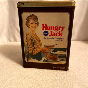 May include: A vintage tin container for Hungry Jack Buttermilk Complete Pancake Mix. The tin is brown with a gold trim and features a woman in a vintage dress making pancakes. The text on the tin reads "Hungry Jack Buttermilk Complete Pancake Mix" and "Pillsbury". The net weight is 32 oz (2 lb) or 907g.