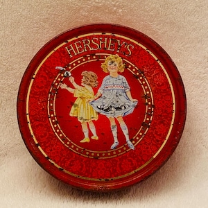 May include: Vintage red tin with a gold border and a floral design. The tin features a picture of two girls in dresses, one wearing a yellow dress and the other wearing a blue dress. The text "HERSHEY'S" is printed in gold letters on the top of the tin.
