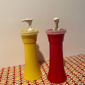 May include: Two vintage condiment dispensers. One is yellow, the other is red. Both have white pump tops and clear lids. The dispensers are on a red and white patterned surface.