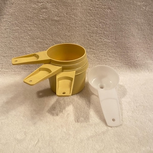 May include: A set of yellow measuring cups stacked together, with handles extending from the sides. A single white measuring cup is next to the stack. The cups are made of plastic and are designed for kitchen use.