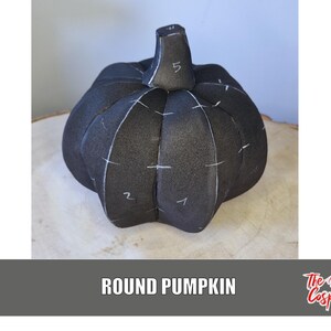 Pode incluir: Uma abóbora preta e redonda com marcas brancas e um caule. A abóbora está sobre uma superfície de madeira castanha clara. O texto "ROUND PUMPKIN" está na parte inferior da imagem.