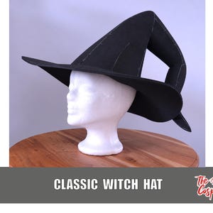 Puede incluir: Un sombrero de bruja negro con una copa alta, puntiaguda y ligeramente curvada, y un ala ancha. El sombrero se muestra sobre una cabeza de maniquí blanca. El texto "CLASSIC WITCH HAT" y "The Dodo Cosplay" son visibles en la parte inferior.