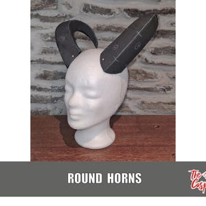 Puede incluir: Un par de cuernos redondos negros hechos de cartón. Los cuernos están unidos a una cabeza de maniquí blanca. El texto "ROUND HORNS" está impreso debajo de la imagen. El texto "The Dodo Cosplay" está impreso en la esquina inferior derecha de la imagen.