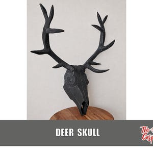 Puede incluir: Un cráneo de ciervo negro para montaje en pared con una construcción geométrica facetada y líneas de costura blancas visibles. El cráneo tiene astas grandes y ramificadas y un hocico puntiagudo, con el texto DEER SKULL y The Dodo Cosplay.