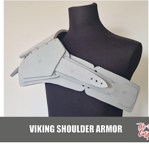 May include: Light gray Viking shoulder armor displayed on a black mannequin. The armor is made of layered, segmented pieces, connected with metal fasteners. The text "VIKING SHOULDER ARMOR" is at the bottom of the image.