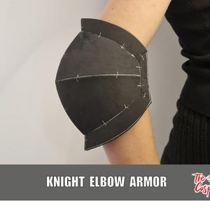 Può includere: Un pezzo di armatura per gomito nero con un design curvo e segmentato, caratterizzato da cuciture bianche visibili lungo le giunture. L'armatura è posizionata su un braccio e il testo KNIGHT ELBOW ARMOR e The Dodo Cosplay sono riportati sotto.