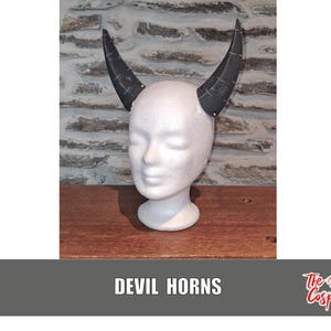 以下が含まれることがあります： 白いマネキンの頭に装着された、黒い発泡スチロール製の悪魔の角のペア。角は湾曲していて先が尖っており、表面はテクスチャ加工されています。角はコスプレやコスチューム用です。