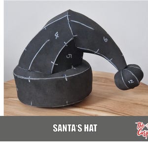 Könnte beinhalten: Ein schwarzer Weihnachtsmannhut mit weißen Markierungen und einem abgerundeten Bommel. Der Hut besteht aus einem dunklen Material und liegt auf einer Holzoberfläche. Der Text "SANTA'S HAT" wird am unteren Bildrand angezeigt.
