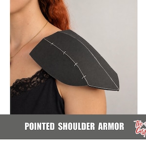 Può includere: Un pezzo di armatura per spalla nero e appuntito con cuciture bianche visibili lungo le giunture. L'armatura è composta da più pannelli uniti insieme, creando una forma angolare e strutturata che poggia sulla zona della spalla.