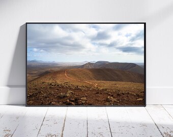 May include: A framed landscape photograph featuring a winding dirt road across a series of brown hills under a cloudy sky. The photograph is displayed on a white wall above a white wooden floor.