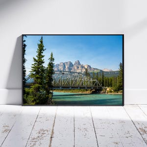 May include: Framed landscape photograph featuring a steel bridge spanning a turquoise river, with evergreen trees and a mountain range under a clear blue sky. The bridge has a lattice design.