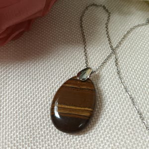 Puede incluir: Un collar con colgante que presenta una piedra de ojo de tigre pulida en forma de lágrima. La piedra muestra bandas de tonos marrones y dorados. El colgante está suspendido de una delicada cadena de color plateado, creando un diseño simple y elegante.