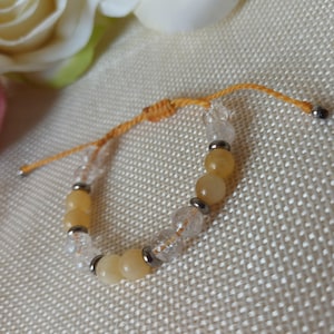 Peut inclure: Bracelet réglable avec des perles jaunes et transparentes alternées. Les perles jaunes sont rondes et opaques, tandis que les perles transparentes sont facettées. Des accents métalliques argentés séparent les perles. Le bracelet est monté sur un cordon jaune.