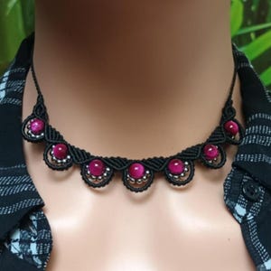 Op de afbeelding: Een zwarte macramé ketting met helder roze edelstenen gezet in decoratieve zilverkleurige zettingen. De ketting heeft een geschulpte patroon, waardoor een uniek en opvallend accessoire ontstaat. De ketting is een choker stijl.