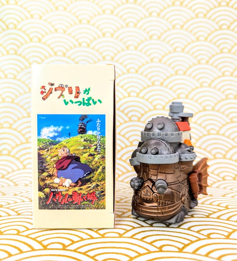 Peut inclure: Une figurine de style steampunk de couleur bronze avec un design d&eacute;taill&eacute;, ressemblant &agrave; un v&eacute;hicule ou une machine. &Agrave; c&ocirc;t&eacute;, une bo&icirc;te avec du texte japonais et une illustration d'une sc&egrave;ne de film. Le fond a un motif de vagues or et blanc.