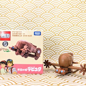 May include: A brown and tan toy airplane with a propeller and a skull and crossbones design. The airplane is from the movie "Castle in the Sky" and is part of the "Dream Tomica" series. The box is labeled "Ghibli Collection" and "Tiger Moth".
