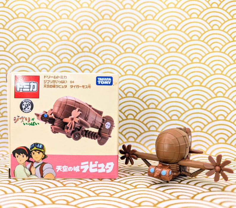 May include: A brown and tan toy airplane with a propeller and a skull and crossbones design. The airplane is from the movie "Castle in the Sky" and is part of the "Dream Tomica" series. The box is labeled "Ghibli Collection" and "Tiger Moth".