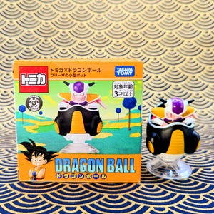 Könnte beinhalten: Eine Dragon Ball Z Frieza-Figur und ihre Box. Die Figur ist schwarz, orange und lila. Die Box ist orange und grün mit japanischem Text und einem Bild von Goku. Auf der Box steht "Dragon Ball".