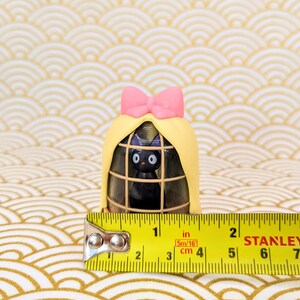 Op de afbeelding: Een klein zwart kattenbeeldje met een roze strik zit in een gele kooi met een bruine houten basis. De kooi is 9 cm hoog.