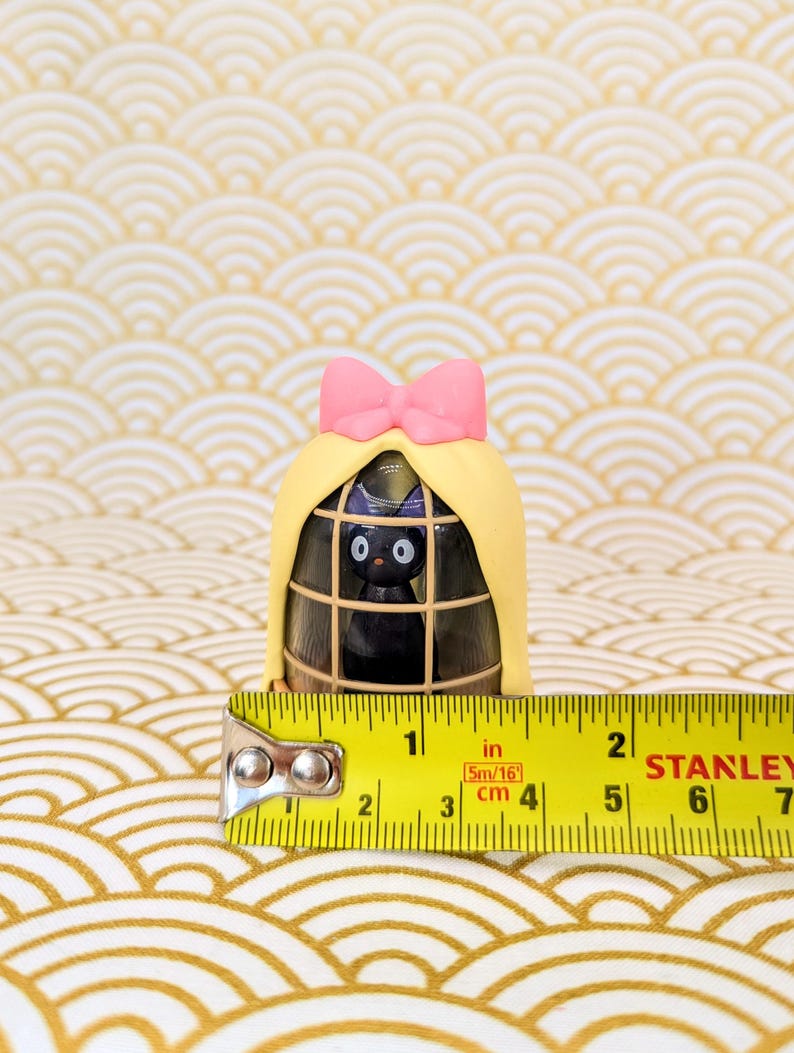 Op de afbeelding: Een klein zwart kattenbeeldje met een roze strik zit in een gele kooi met een bruine houten basis. De kooi is 9 cm hoog.