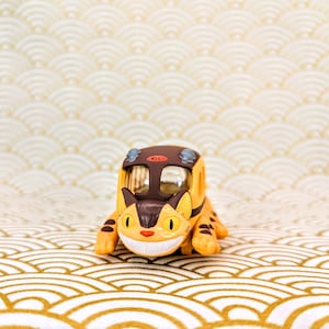 May include: A yellow and brown toy train shaped like a cat with a smiling face. The train is on a white and gold patterned surface.