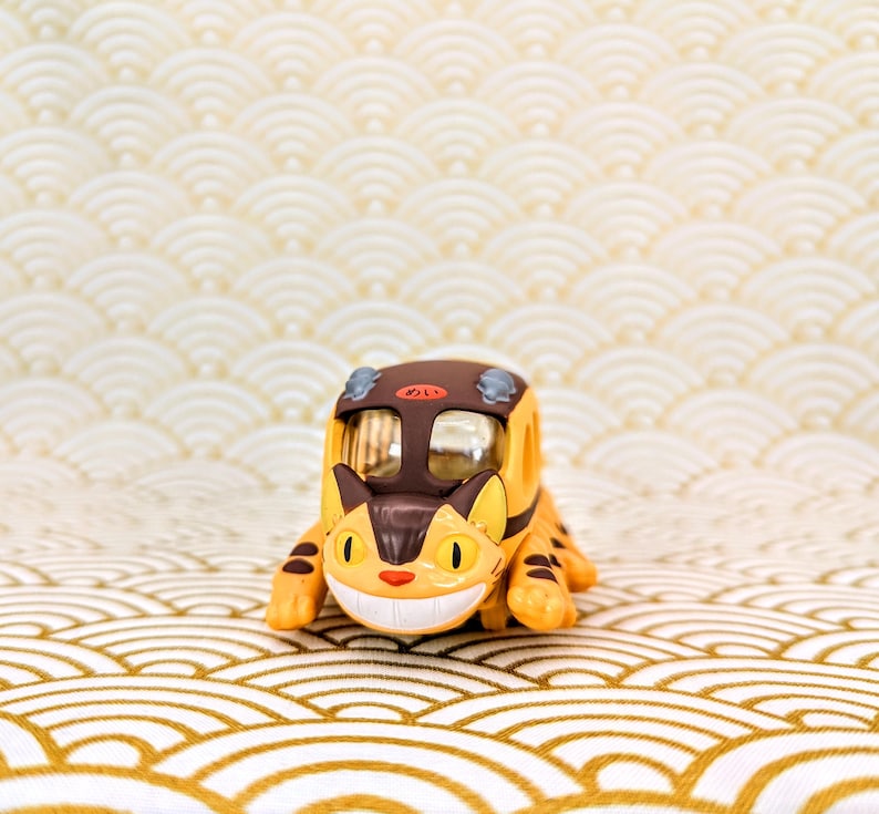 May include: A yellow and brown toy train shaped like a cat with a smiling face. The train is on a white and gold patterned surface.