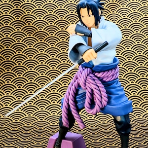 Pode incluir: Estatueta de um personagem em posi&ccedil;&atilde;o de combate, segurando uma espada e um objeto preto. A figura usa uma roupa branca e azul com um cinto de corda roxa. O fundo &eacute; um design dourado e preto.