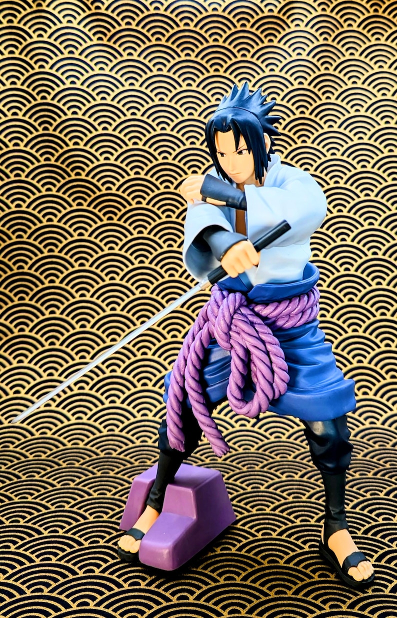 Pode incluir: Estatueta de um personagem em posi&ccedil;&atilde;o de combate, segurando uma espada e um objeto preto. A figura usa uma roupa branca e azul com um cinto de corda roxa. O fundo &eacute; um design dourado e preto.