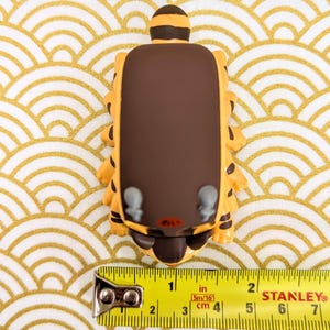 May include: A brown and yellow cartoon animal figurine with a brown back and a yellow face. The figurine is about 2 inches tall and is sitting on a white and gold patterned surface.