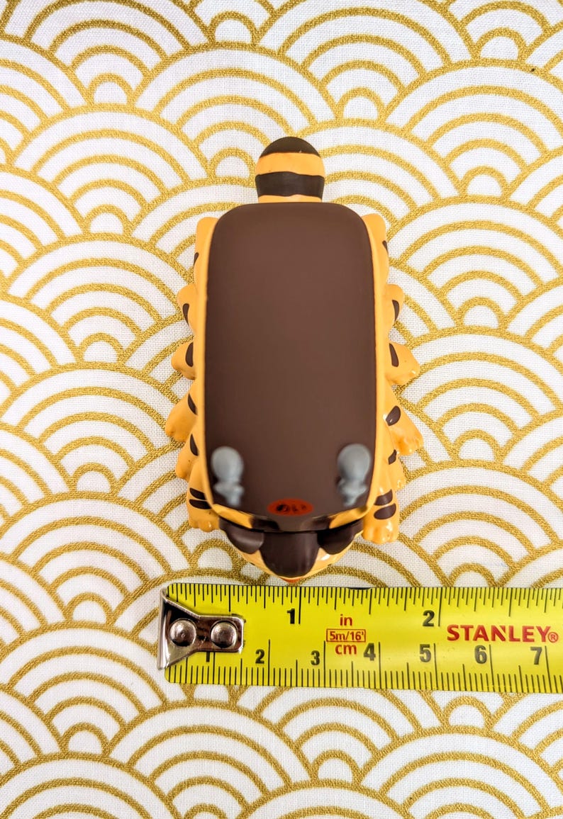 May include: A brown and yellow cartoon animal figurine with a brown back and a yellow face. The figurine is about 2 inches tall and is sitting on a white and gold patterned surface.