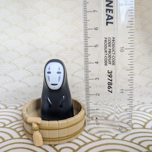 May include: A black No Face character from the animated film Spirited Away, sitting in a small wooden tub with a wooden paddle. The character has a white face with a purple mouth and eyes.