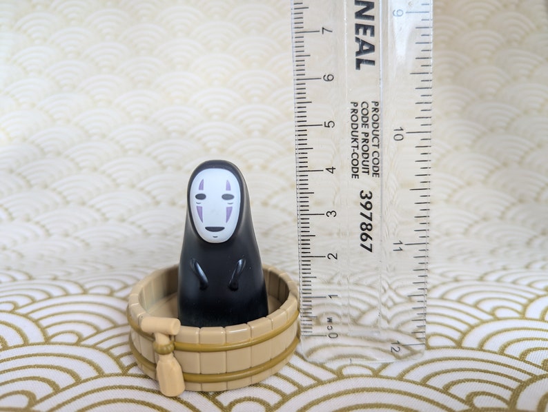 May include: A black No Face character from the animated film Spirited Away, sitting in a small wooden tub with a wooden paddle. The character has a white face with a purple mouth and eyes.