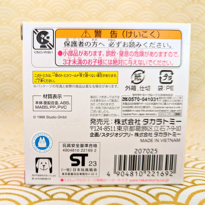 May include: A white cardboard box with a warning label about small parts and a choking hazard. The box features a red and white warning symbol, a picture of a Totoro character, and the text "Studio Ghibli". The box also includes a barcode, a product code, and the text "Made in Vietnam".
