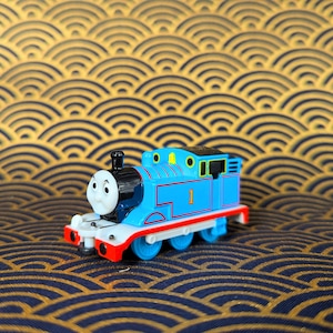 May include: A blue toy train engine with a black face and yellow eyes. The engine has a number "1" on the side and is on a gold and black patterned surface.