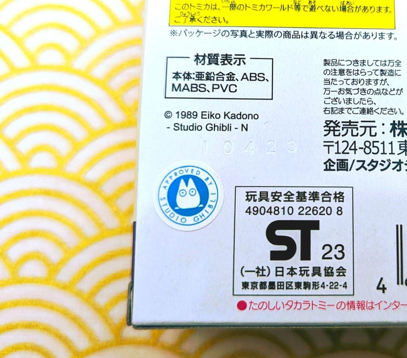 Op de afbeelding: Een witte doos met een geel en wit patroon aan de linkerkant. De doos heeft een blauwe en witte sticker met een cartoonkat erop. De doos heeft ook een witte sticker met zwarte tekst die "Approved by Studio Ghibli" zegt. De doos heeft zwarte tekst die "Materiaal: Zinklegering, ABS, MABS, PVC" zegt. De doos heeft zwarte tekst die "1989 Eiko Kadono - Studio Ghibli - N" zegt. De doos heeft zwarte tekst die "10423" zegt. De doos heeft zwarte tekst die "Speelgoed Veiligheidsstandaard Goedgekeurd 4904810 22620 8" zegt. De doos heeft zwarte tekst die "ST 23" zegt. De doos heeft zwarte tekst die "(Vereniging) Japanse Speelgoedvereniging Tokyo Sumida-ku Higashikomagata 4-22-4" zegt. De doos heeft zwarte tekst die "4" zegt. De doos heeft zwarte tekst die "Leuke Tsum Tsum informatie is op internet beschikbaar" zegt.