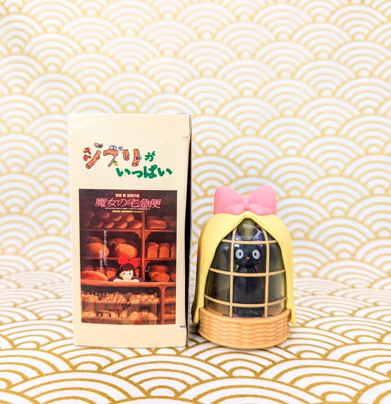 Op de afbeelding: Een klein, geel en roze beeldje van een zwarte kat in een kooi. Het beeldje is uit de film "Kiki's Bezorgservice" en heeft de Japanse tekst "魔女の宅急便" wat zich vertaalt naar "Kiki's Bezorgservice".