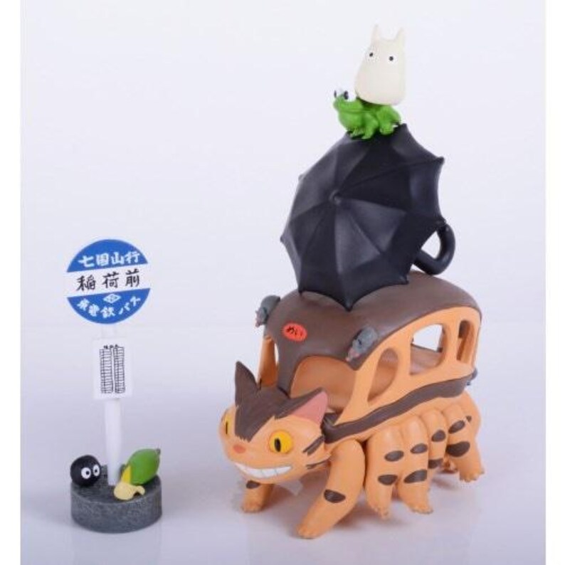 May include: A whimsical figurine featuring a cat-bus with an umbrella, a Totoro figure, and a green frog. A sign with Japanese text is also included. The cat-bus is orange and brown, with a smiling face.