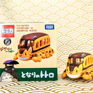 May include: A yellow and brown toy bus with cat ears and a smiling face. The bus is on a white background with a box behind it. The box has Japanese text on it that says "Totoro Bus".
