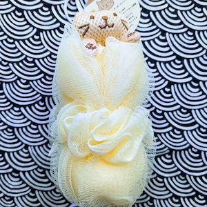 May include: A yellow bath pouf with a small embroidered cat on top. The pouf is on a black and white background with a wave pattern.