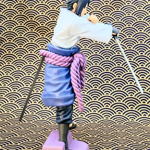 Pode incluir: Uma estatueta de um personagem com cabelo preto espetado, vestindo uma parte superior branca, cal&ccedil;as azuis e um cinto de corda roxa. O personagem est&aacute; segurando uma espada. A estatueta est&aacute; em uma base roxa. O fundo &eacute; um padr&atilde;o de ondas douradas e pretas.